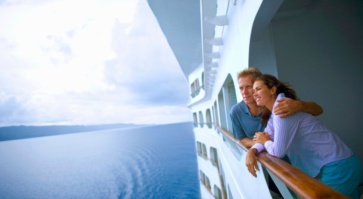 A loving couple looking over the balcony on a cruise ship with smooth blue water and partly cloudy skies