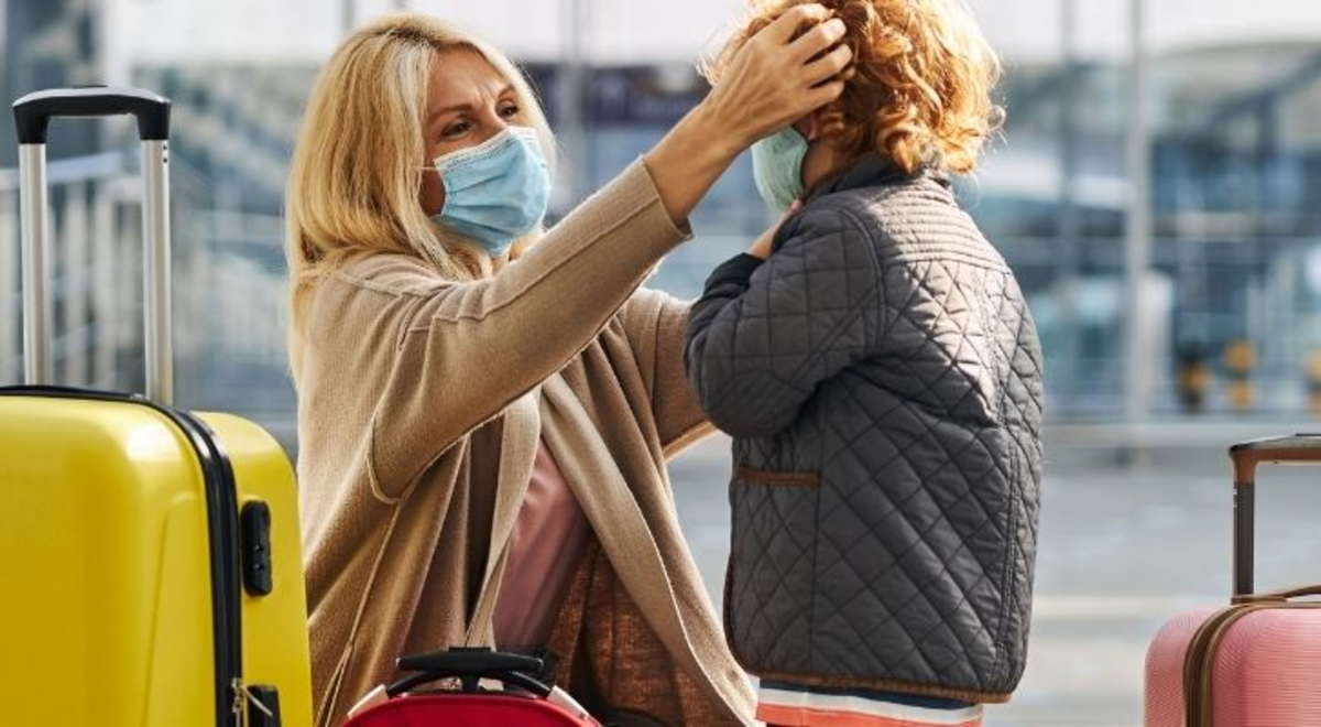 Woman wearing a mask adjusting a child's mask in an airport while another child wearing a mask sits next to her suitcase