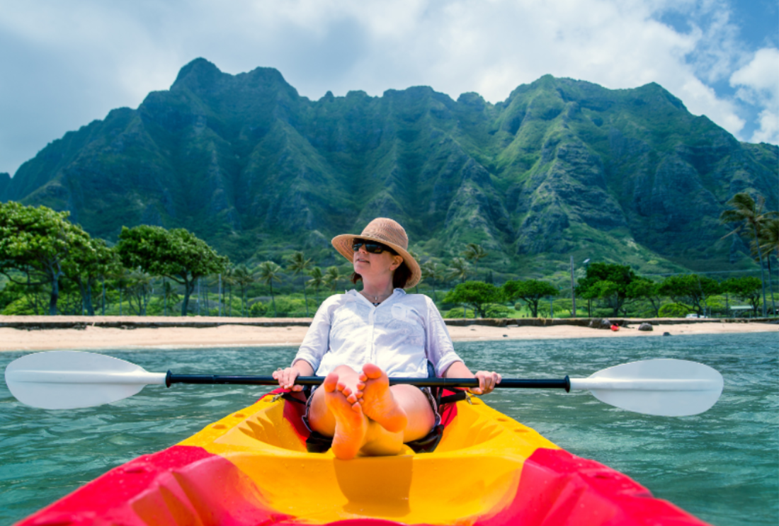 A woman kayaks off the coast Of Hawaii with a forested volcanic range in the background