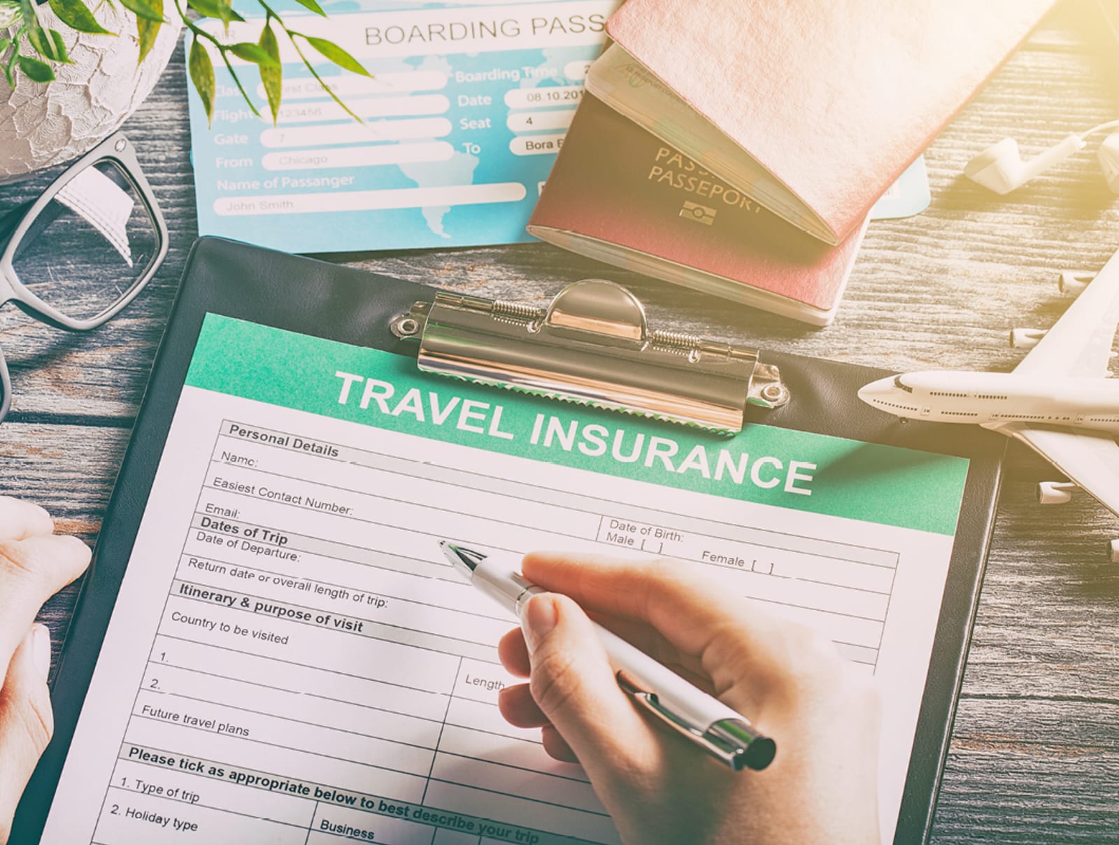 Person's hand filling out travel insurance forms with travel documentation and boarding pass on desk