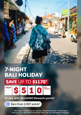 Woman strolling a sunlit Bali market street lined with colourful shops for a 7-night Bali holiday saving up to 1170 from 510 per person