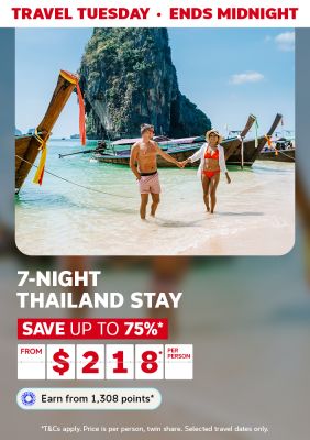 Couple holding hands walking along a Thailand beach past traditional longtail boats for a Travel Tuesday 7-night stay from $218 per person