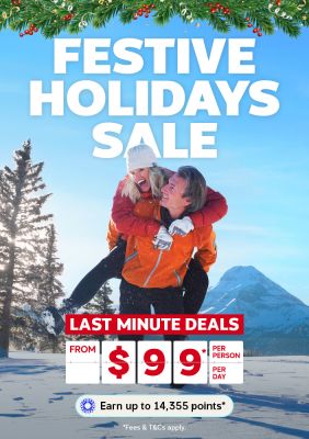 Couple playing in the snow framed by pine trees and snow covered mountains featuring a festive holidays sale with last minute deals from $99 per person per day