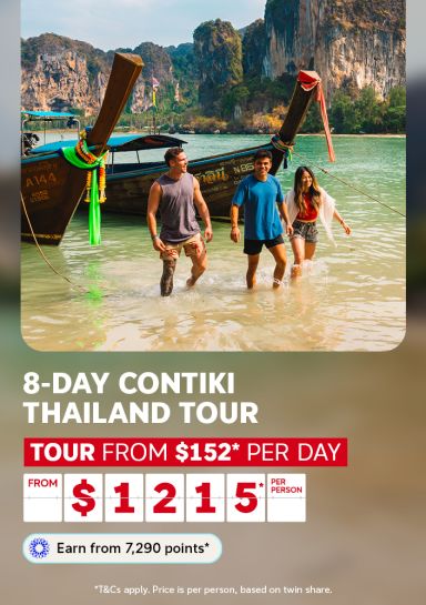 Friends wading ashore from a longtail boat on a Thai beach with dramatic limestone cliffs for an 8-day Contiki Thailand tour from $1215 per person