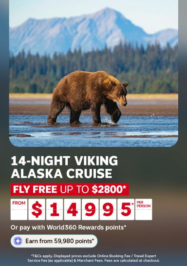 Brown bear wading through water beneath forested mountains for a 14 night Viking Alaska cruise flying free saving up to 2800 from 14995 per person