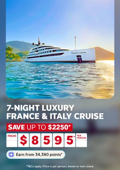 A travel promotional banner featuring the luxury cruise ship Emerald Sakara sailing on calm, clear turquoise water with green hills and a golden sunset in the background. Text reads: "7-Night Luxury France & Italy Cruise – Save up to $2,250* – From $8,595* per person. Earn from 34,380 points*." Fine print states: "T&Cs apply. Price is per person, based on twin share."