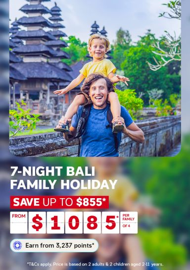 A travel promotional banner featuring a smiling man in a blue t-shirt carrying a young child on his shoulders in front of a traditional Balinese temple. The child, wearing a yellow shirt, smiles and spreads their arms wide. Text below reads: "7-Night Bali Family Holiday – Save up to $855* – From $1,085* per family of 4. Earn from 3,237 points*." Fine print states: "T&Cs apply. Price is based on 2 adults & 2 children aged 2–11 years."