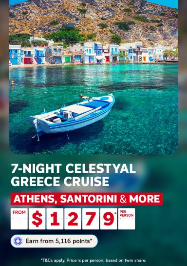 Traditional blue wooden boat moored on crystal clear Greek waters beside colourful Milos village houses for a 7-night Celestyal Greece cruise from $1279