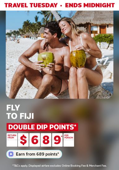 Couple drinking from coconuts on a beach featuring return flights to Fiji from $689* per person