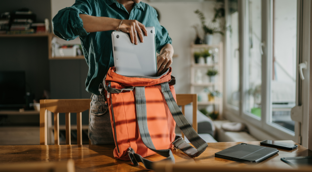 Woma in teal polo shirt putting her laptop in an orange backpack over a wooden table