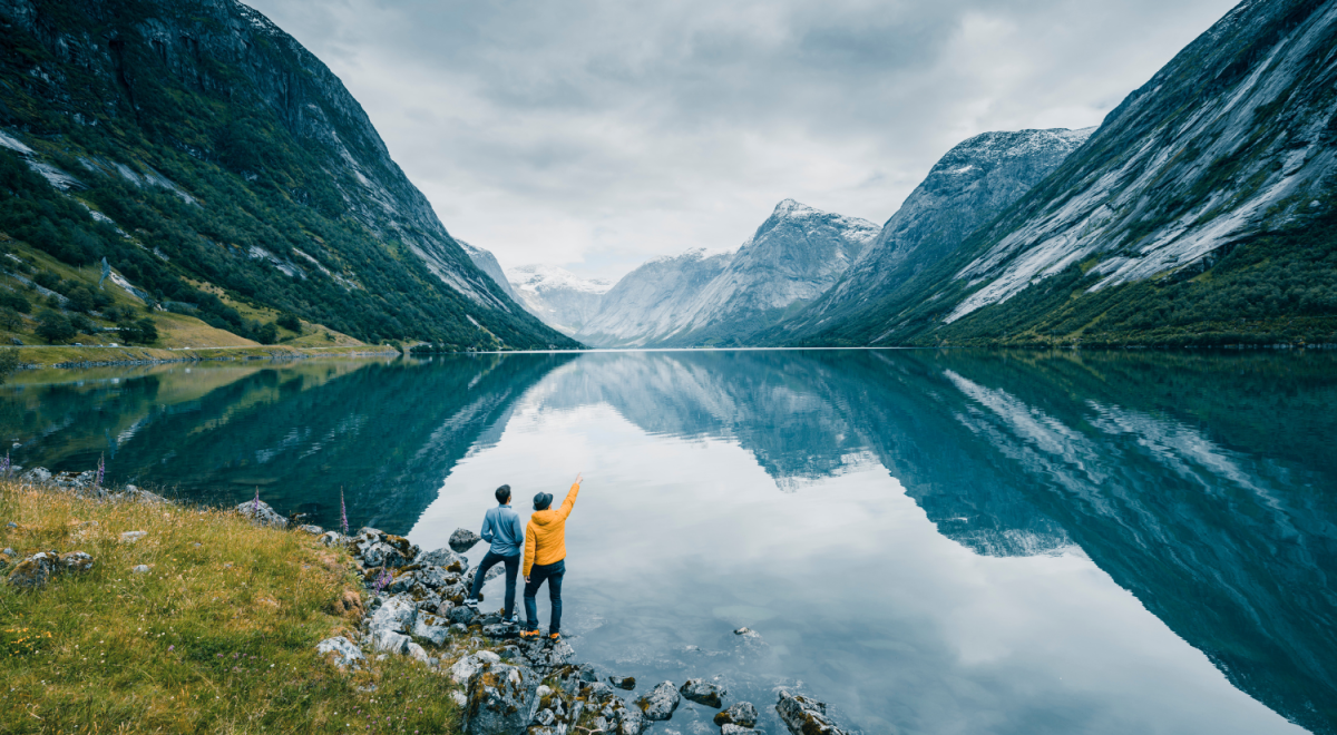Two people stand on rocky ground beside a calm turquoise lake surrounded by steep mountains in a dramatic fjord landscape, with snow-capped peaks in the distance and one person pointing toward the horizon.