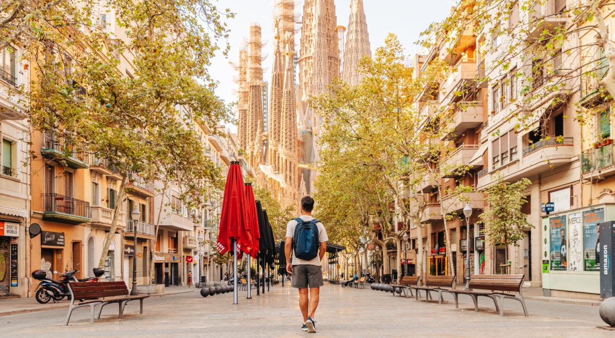 A man is walking down a tree-lined street with his back to the camera. He's wearing a blue backpack. Towering over the background are the intricate spires of Barcelona's Sagrada Família, partially framed by the buildings on either side. Red umbrellas are lined up on the left. The lighting is golden, possibly from a late afternoon sun.