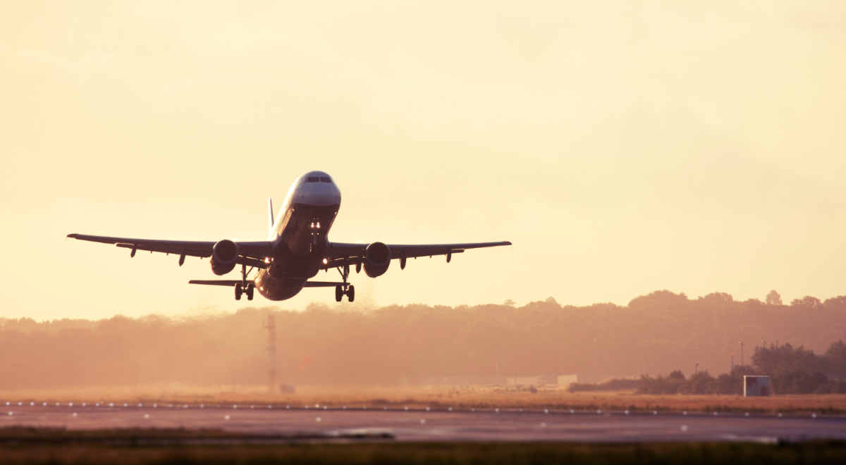 A wide-angle, low-angle shot of a large passenger airplane taking off from a runway during a hazy golden hour. The aircraft is angled upward, positioned slightly left of center, with its landing gear still extended. The sun, positioned behind or to the side of the plane, creates a warm, glowing amber sky and casts the distant treeline and airport structures in a soft, misty silhouette. Bright runway lights are visible in the foreground.