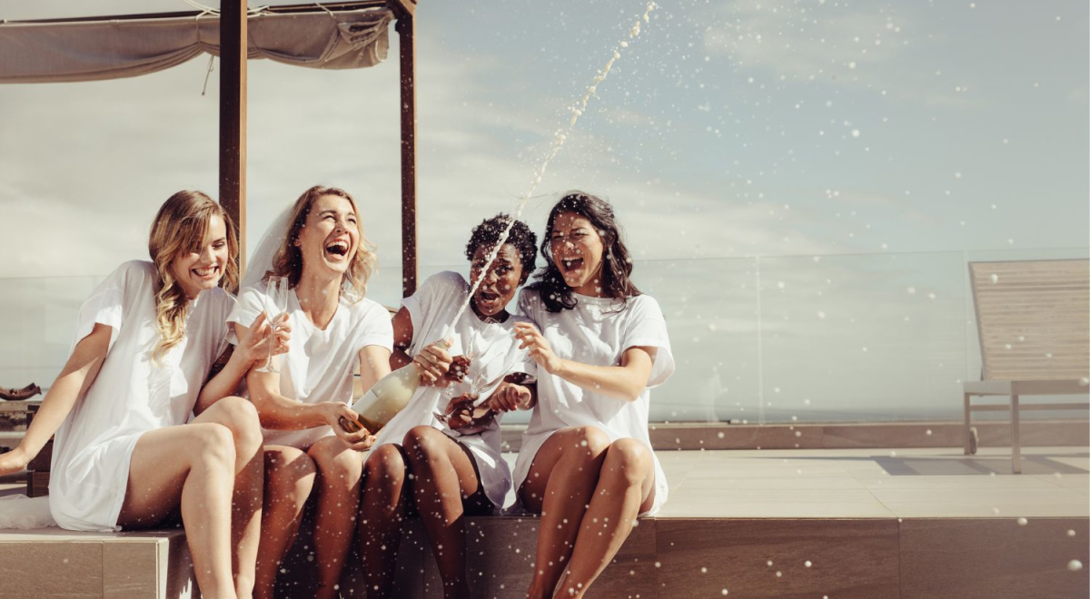 four ladies sitting by a pool popping a bottle of champagne