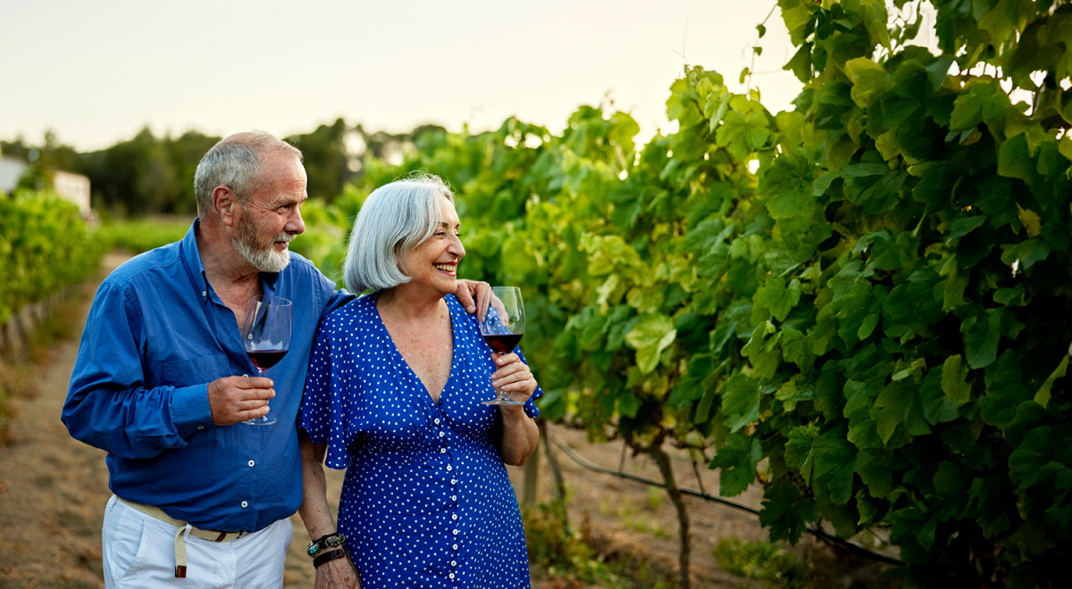 An older couple walking through a vineyard in Europe; they're both holding a glass of red wine