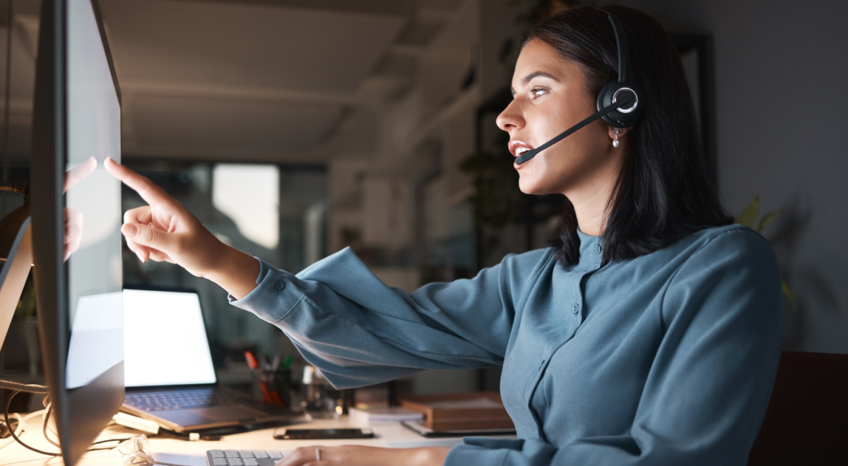 A woman wearing a headset talks while pointing at a computer monitor