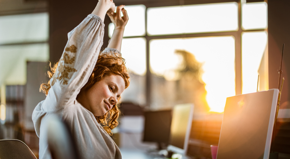 A woman stretches while sitting at her laptop
