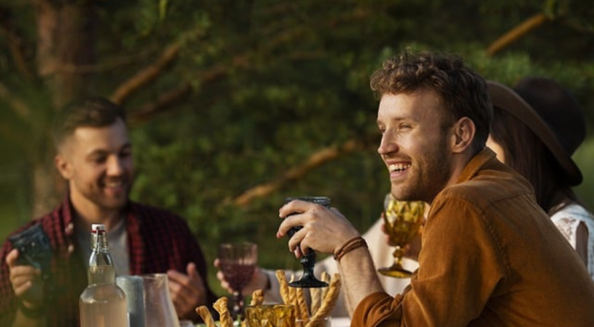 Young man smiles and drinks at the table of a large gathering.
