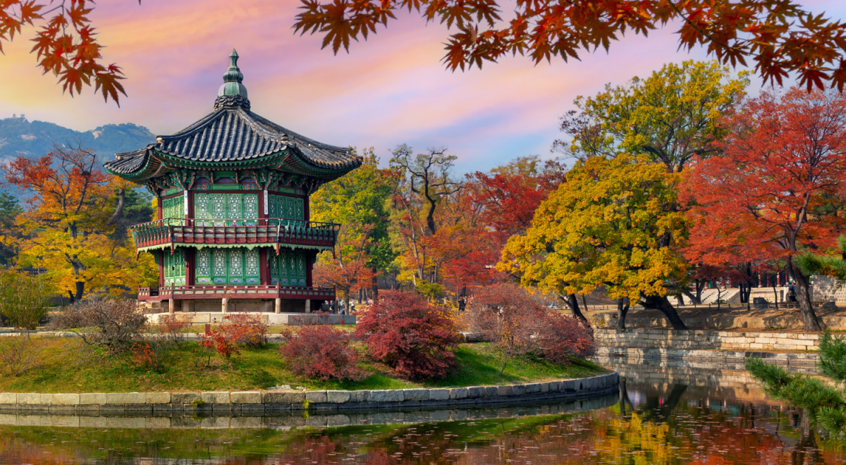The Hyangwonjeong pavilion at Gyeongbokgung Palace in Seoul, surrounded by vivid autumn foliage in red and gold, reflected in a still pond at sunset