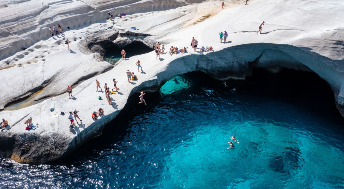 Sandstone cliffs in Greece overhanging clear blue water with people standing near the edge ready to dive into the water