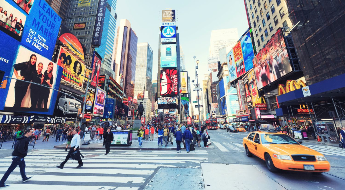 A busy crosswalk in Times Square with people crossing the road and cars driving down the street.