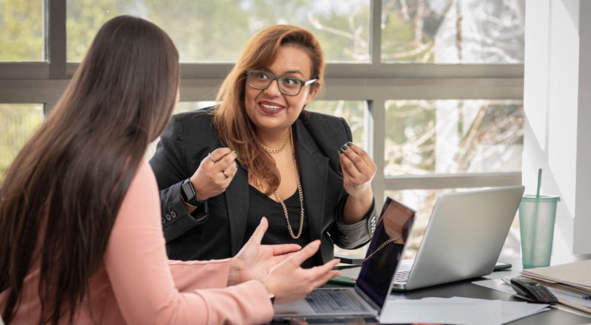 two people talking in an office with laptop on desk between them 