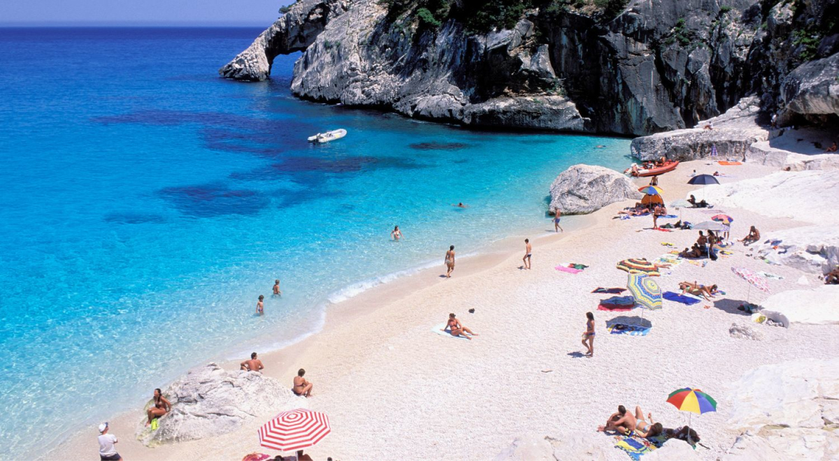 A white sand beach filled with people. Clear blue water with people swimming in it