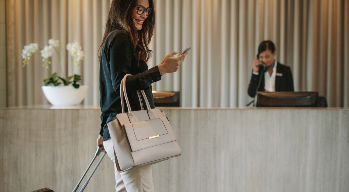 Business lady looking at phone carrying handbag and pulling suitcase walking past a hotel reception desk 