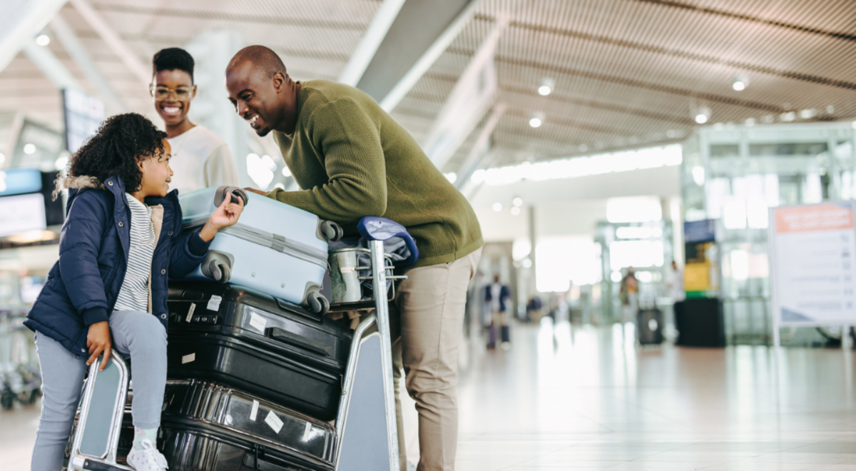 A mother and father looking and smiling at their daughter sitting on luggage on trolley in airport