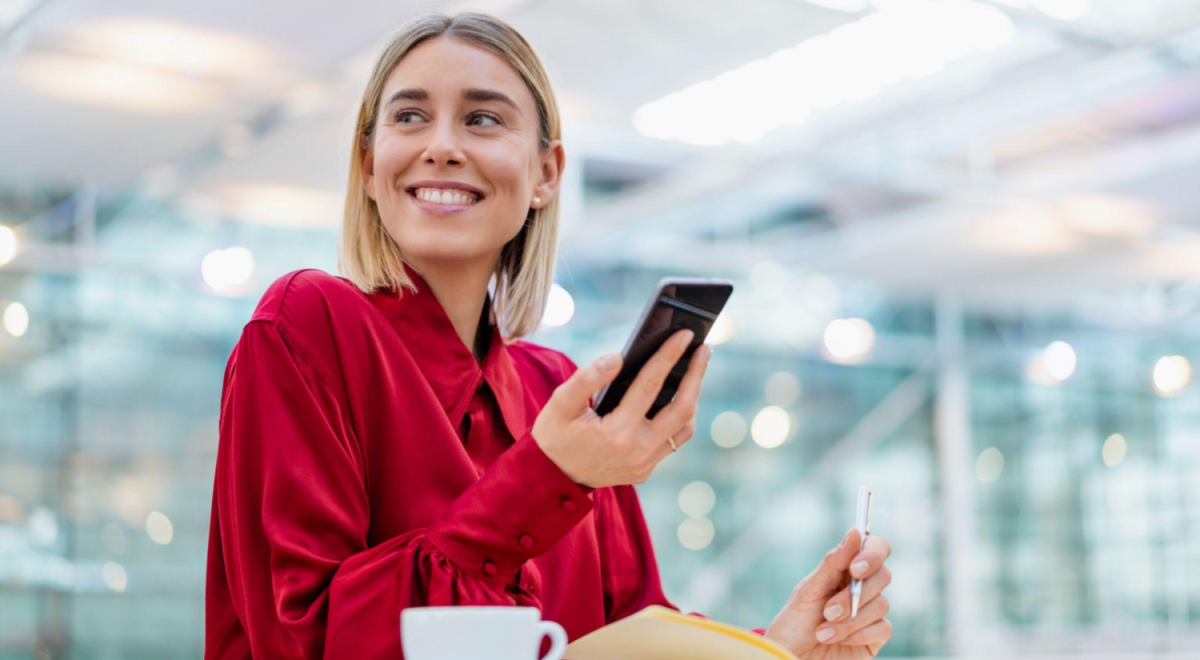 business person in red shirt sits with phone in hand writing in notebook with coffee next to them