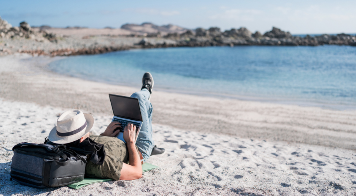 Man lying on beach resting head on backpack while working on his laptop