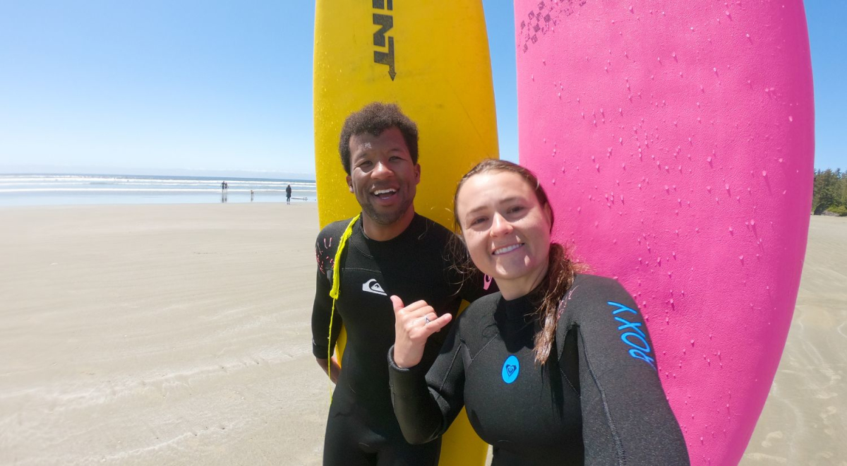 Two people standing on a beach smiling. Behind them is a yellow and pink surfboard stuck in the sand so that it stands up.