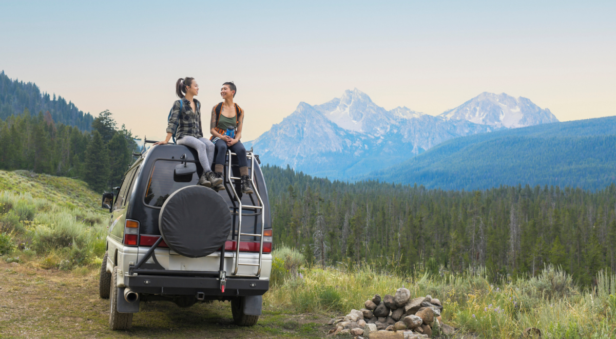 two women sitting on roof of large van with mountains and forest in background