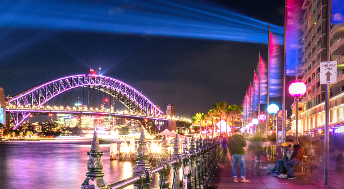 Sydney Harbour Bridge lit in violet with dramatic blue light beams radiating across the harbour waterfront during Vivid Sydney at night