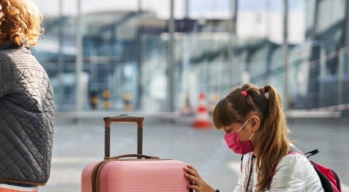 Woman wearing a mask adjusting a child's mask in an airport while another child wearing a mask sits next to her suitcase