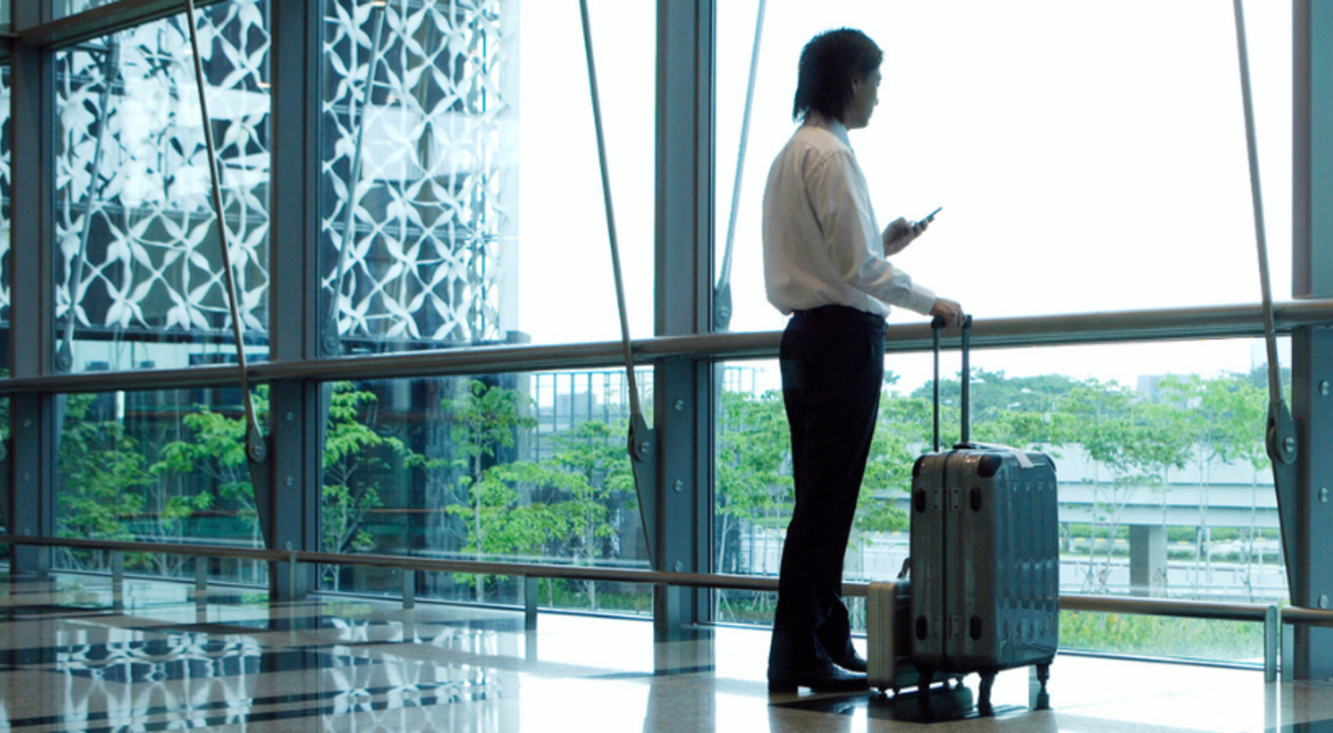 an asian guy facing the windows on the airport while looking on his phone and holding his luggage on his other hand