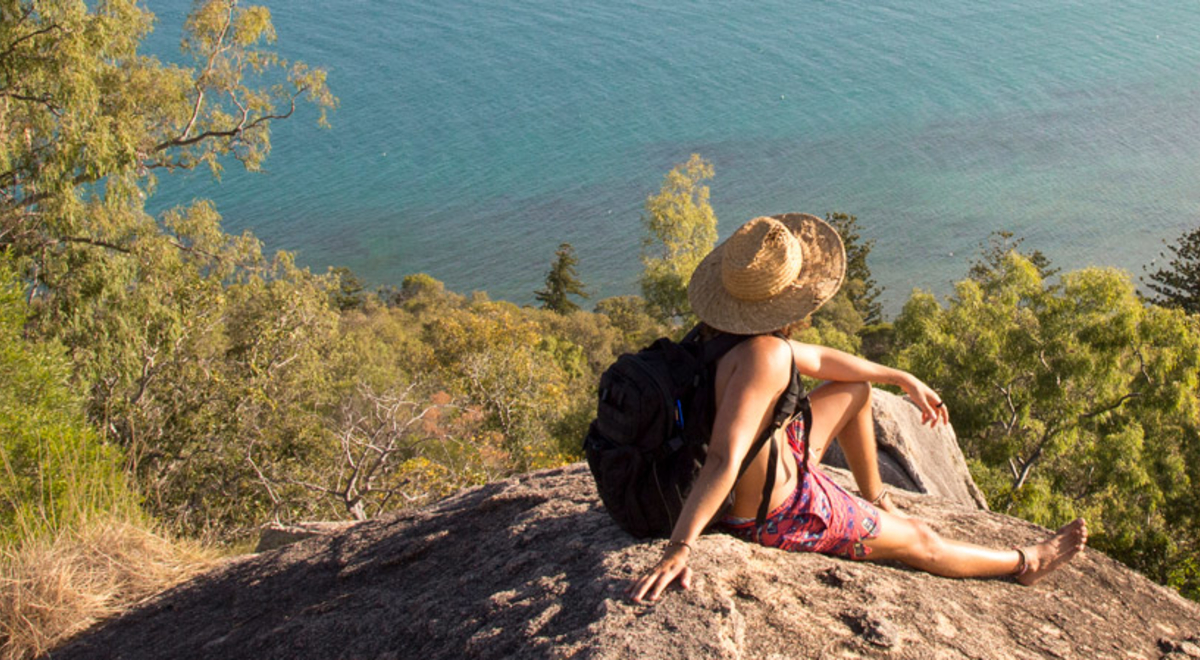 A guys sitting at the edge of stone on top of the mountain looking at the sea