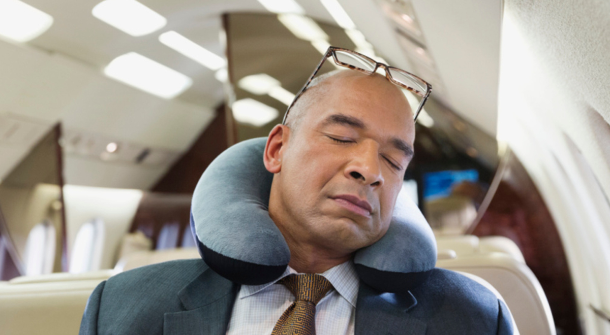 A man wearing a suite sleeping on a plane with a neck pillow around his neck