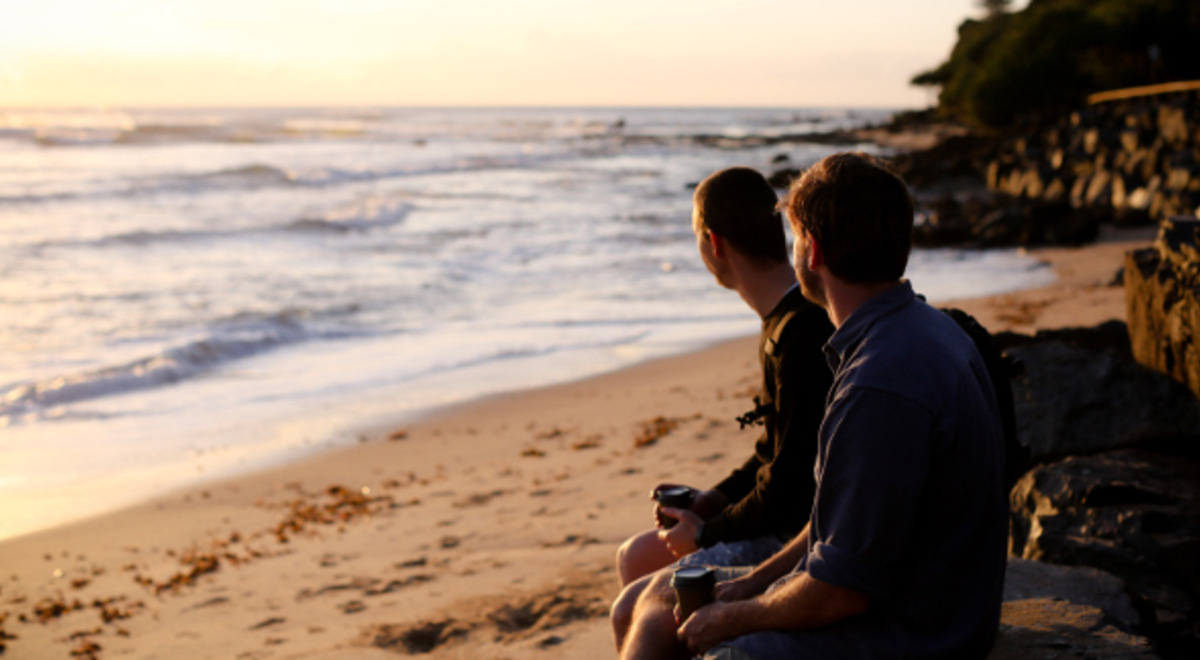 Two gentlemen sitting on a rock enjoying a cup of coffee while gazing out into the sea