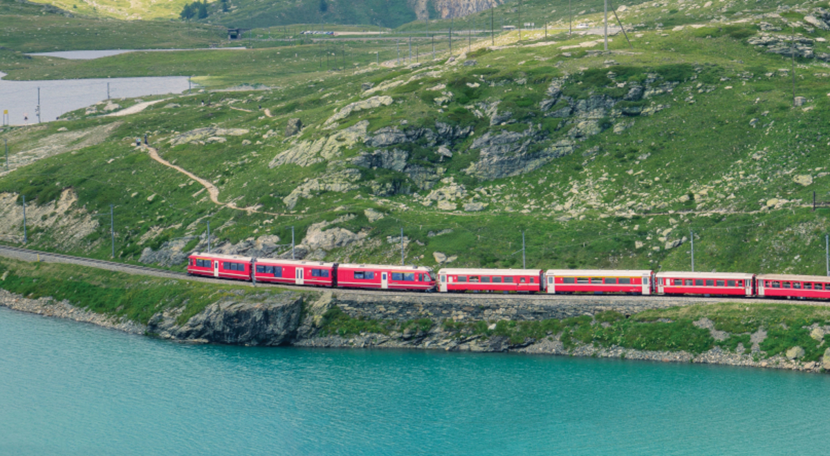 Red train going past a lake and green mountains