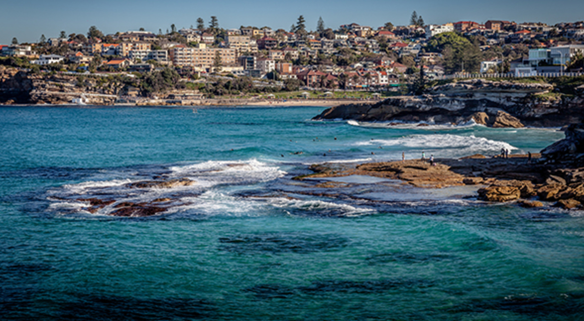 Ocean that is part of the Bondi to Cogee walk. Rocks and waves are shown with buildings in the background. 