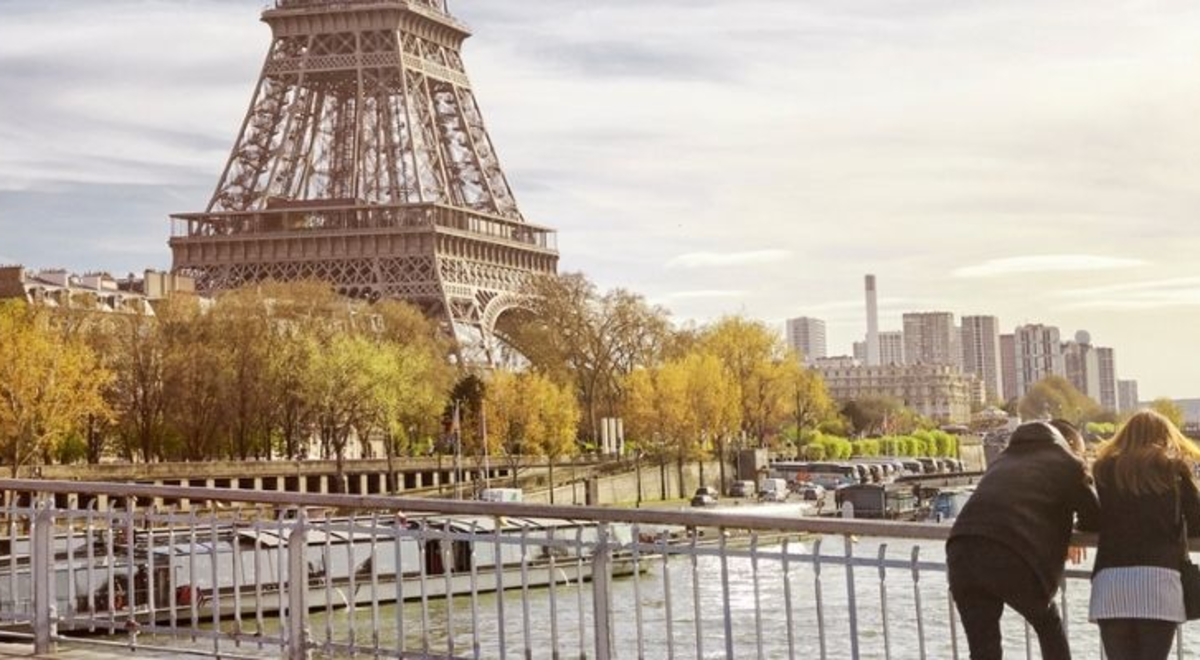 Femme and masculine presenting people stand on bridge over looking Seine River in Paris. the Eiffell Tower is in the background. 