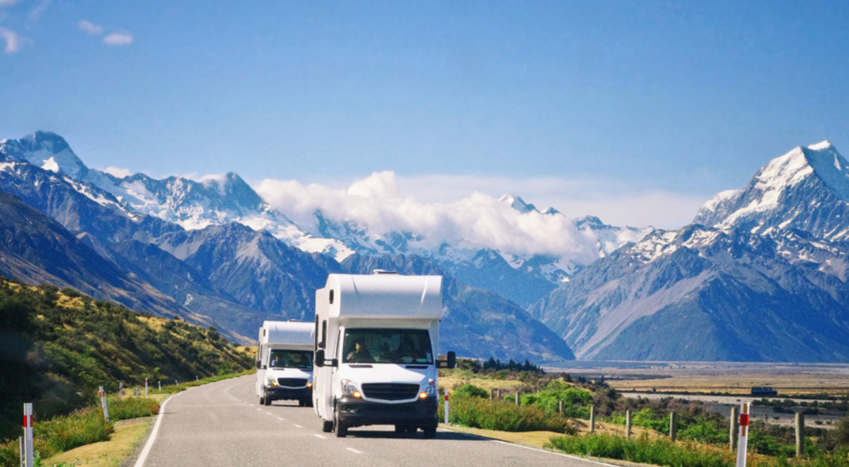 Two campervan head down a straight road with the snowy Southern Alps and Mount Cook in the background on a clear day.