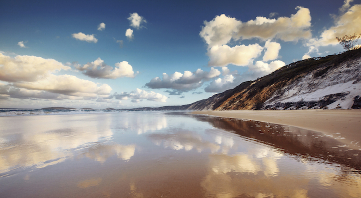 Clouds in a blue sky are reflected in still shallow on a wide beach by sand cliffs at Rainbow Beach, Queensland