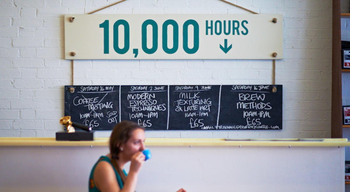 A woman sits at a table drinking from a blue coffee cup beside a counter and menu in a London cafe blackboard 