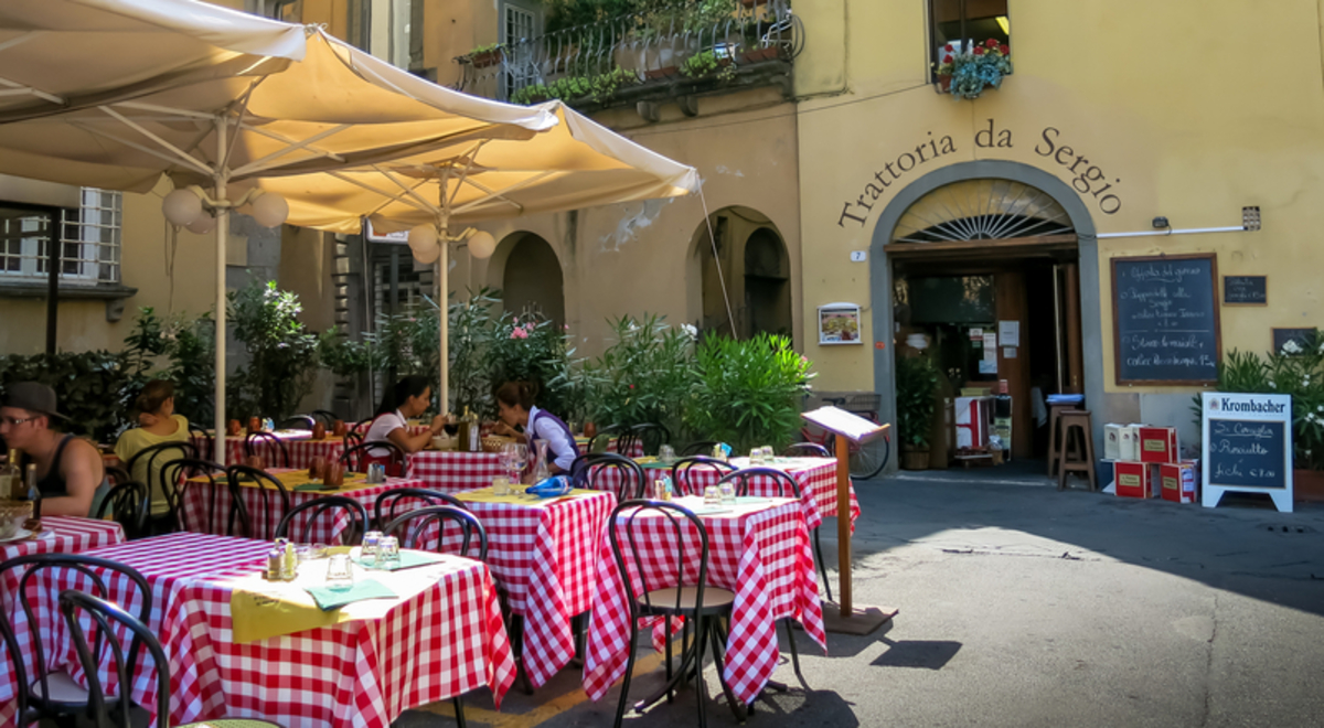 People sit at tables with red-and-white checked tablecloths at an el fresco cafe in front of a large Romanesque building with tall archways.
