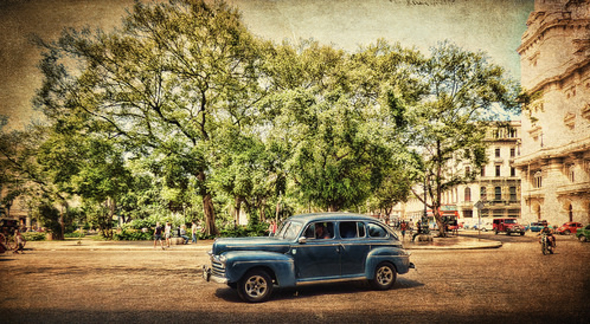 A vintage blue car drives past a large tree and buildings