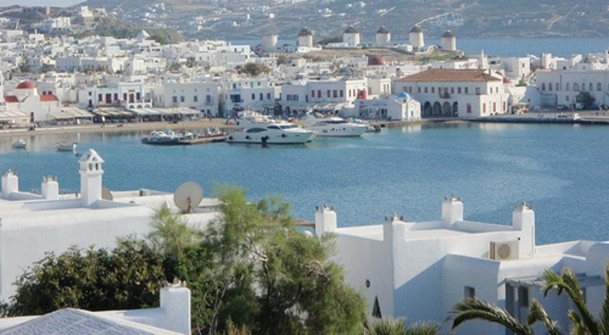 Luxury leisure boats moored at a marina by a hilly seaside town with stucco buildings.
