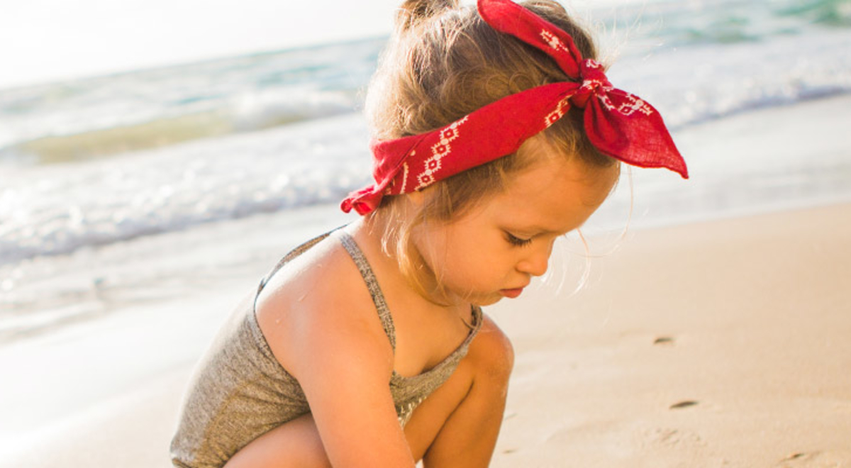 Young girl crouching on the beach wearing a grey swimsuit and a red bandanna 