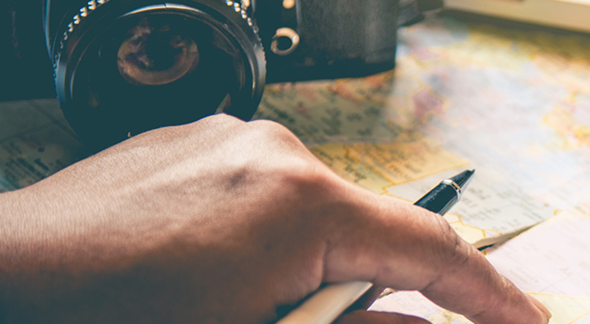 A man's hand holding a pen, a camera, sunglasses and a summer hat rest on an open atlas